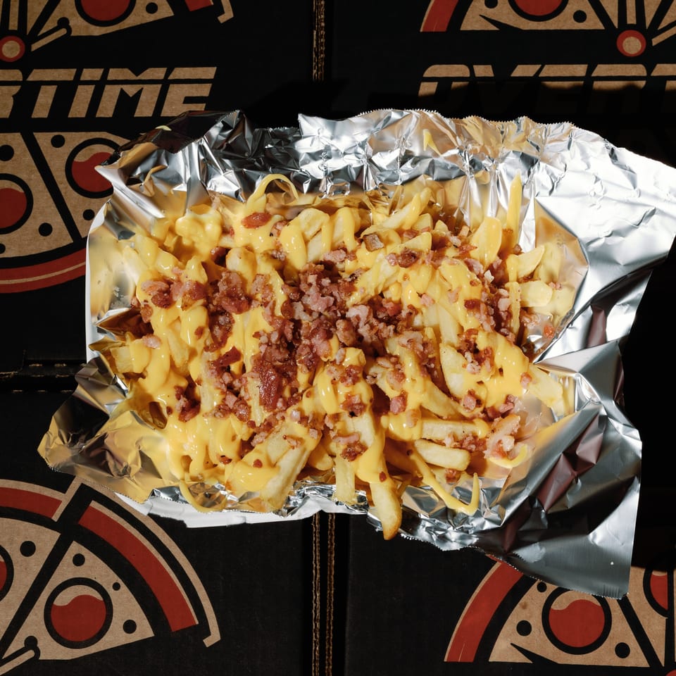Cheesy fries topped with bacon bits in a foil container, on an Overtime Pizza box.