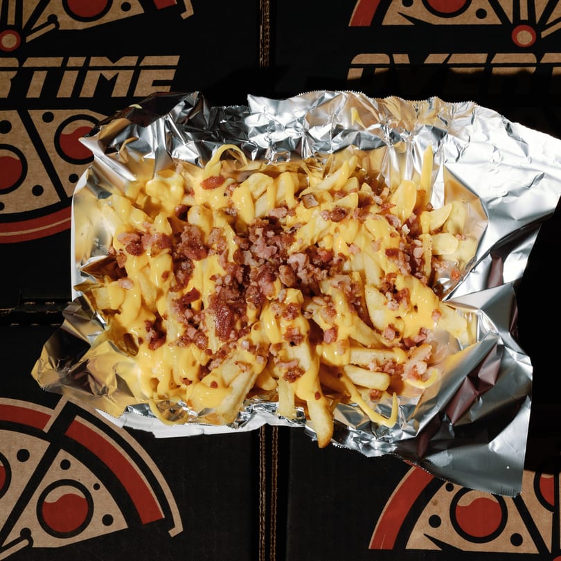 Cheesy fries topped with bacon bits in a foil container, on an Overtime Pizza box.