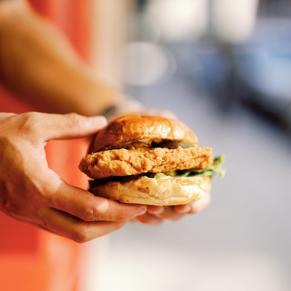 Hands holding a crispy fried chicken sandwich on a brioche bun with lettuce and sauce.